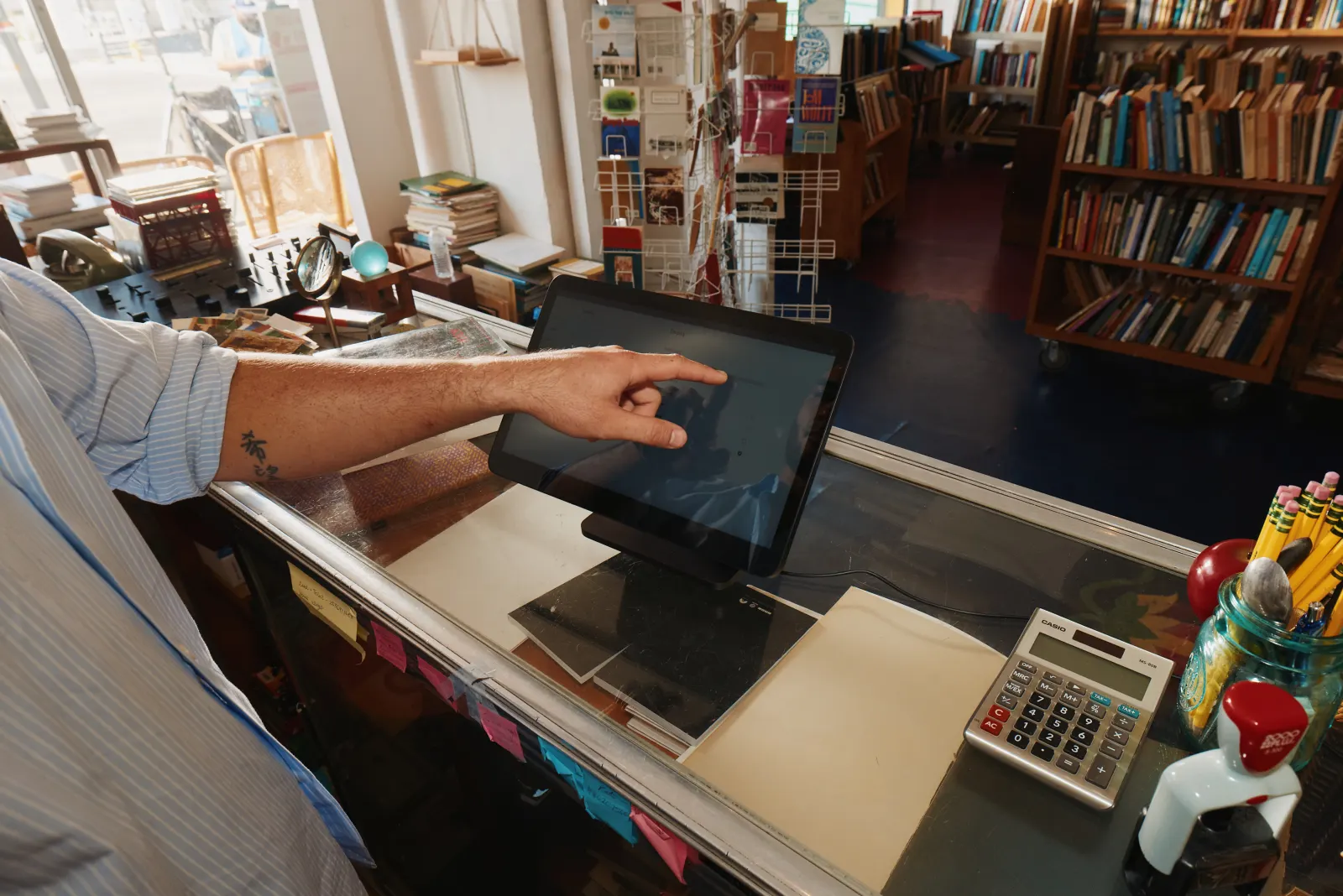 A bookshop counter, where a seller reaches across to a Square register beside a
             calculator and a jar of pencils.
